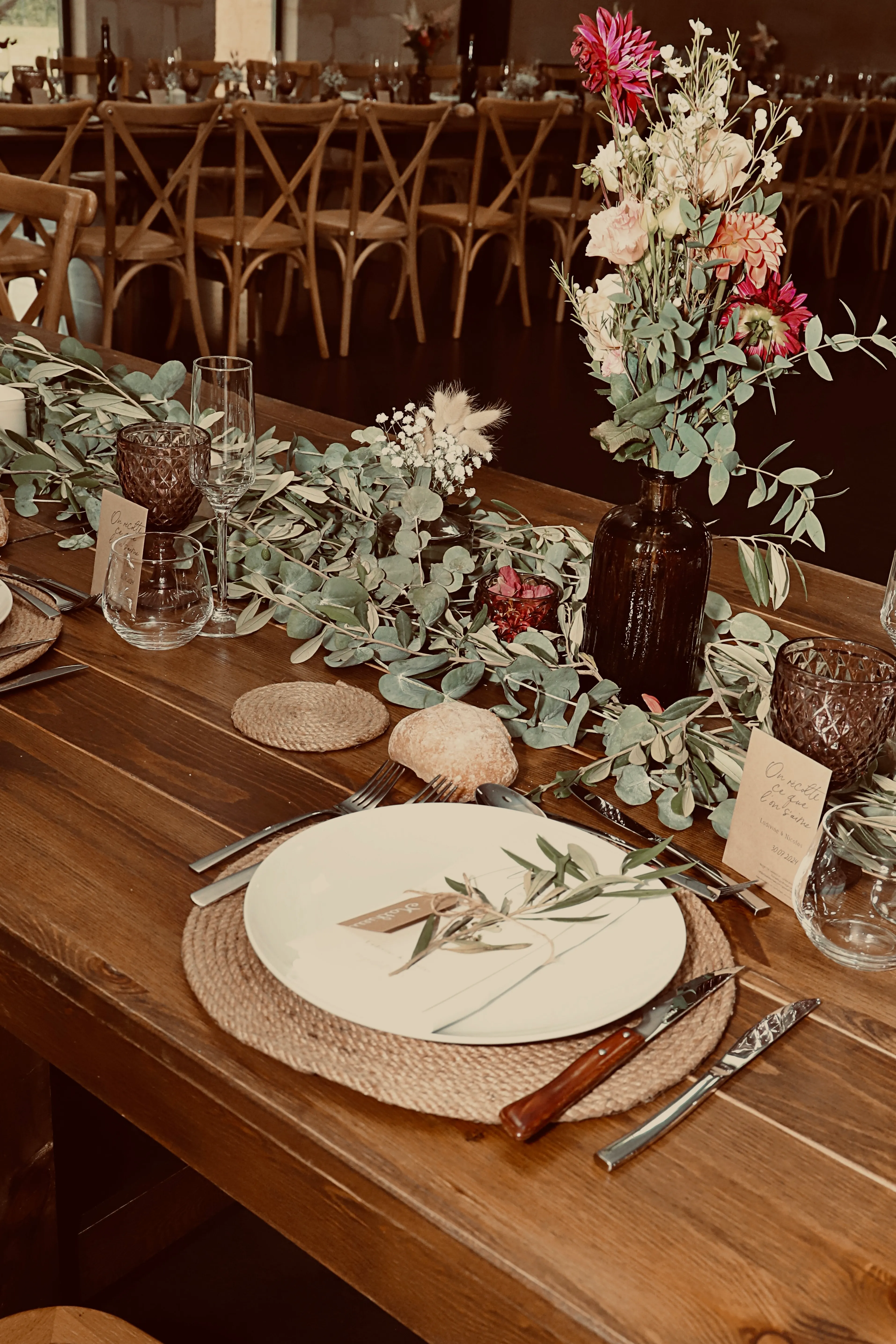 Une longue table de réception dressée : chemin de table en eucalyptus, bonbonne de fleurs, vaisselle et bougies.